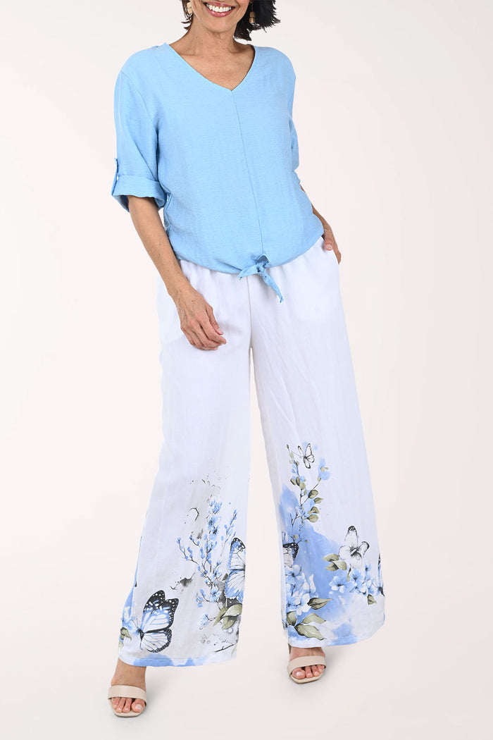 Woman wearing a light blue top and white pants with floral patterns on a plain background